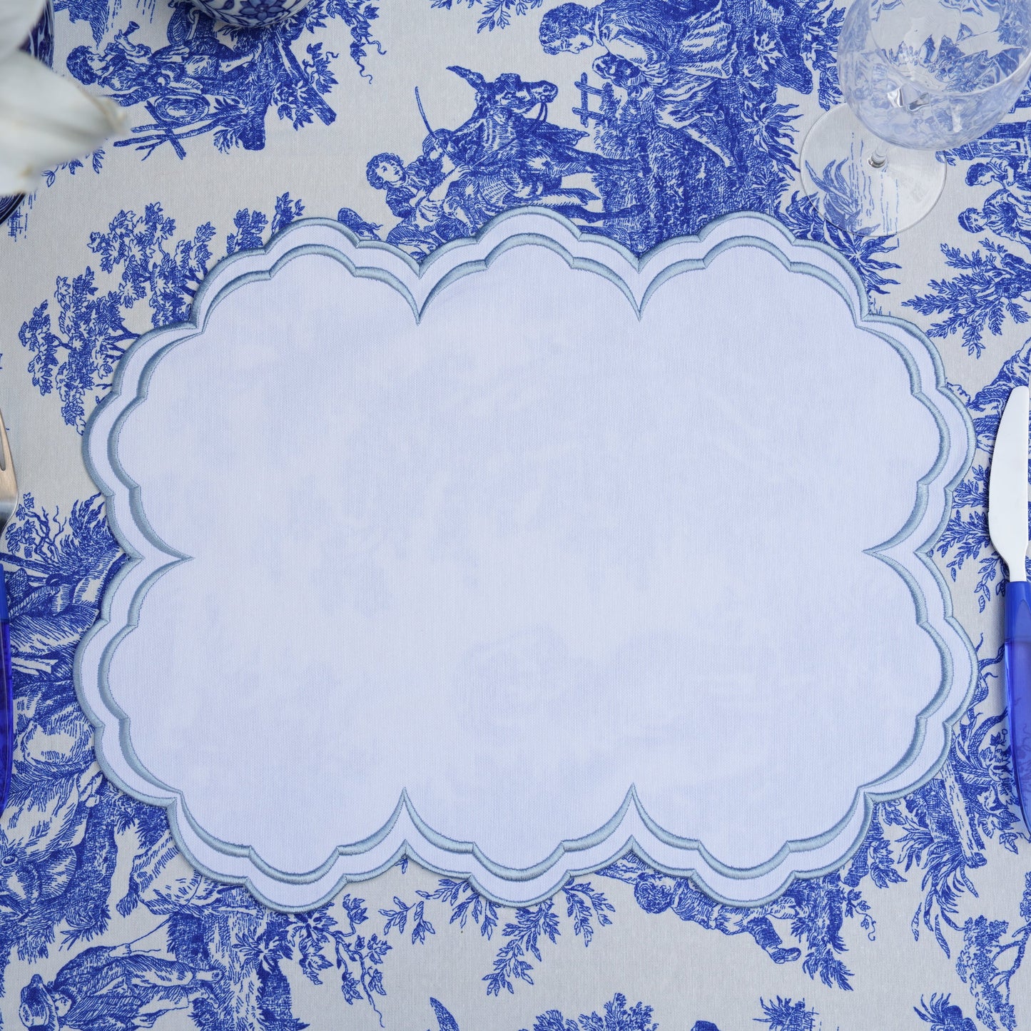 A blue and white floral patterned placemat with a scalloped edge, laid out on a table with cutlery at either side.