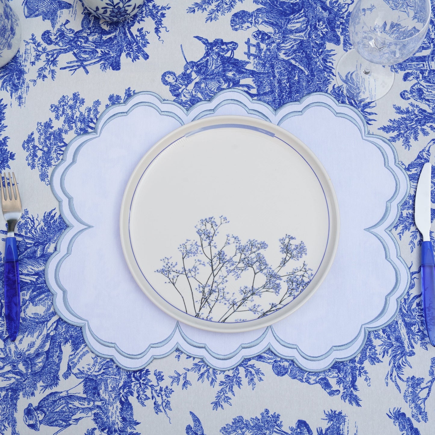 A blue and white floral patterned placemat with a round white plate and blue cutlery on top of it, displayed against a white background.