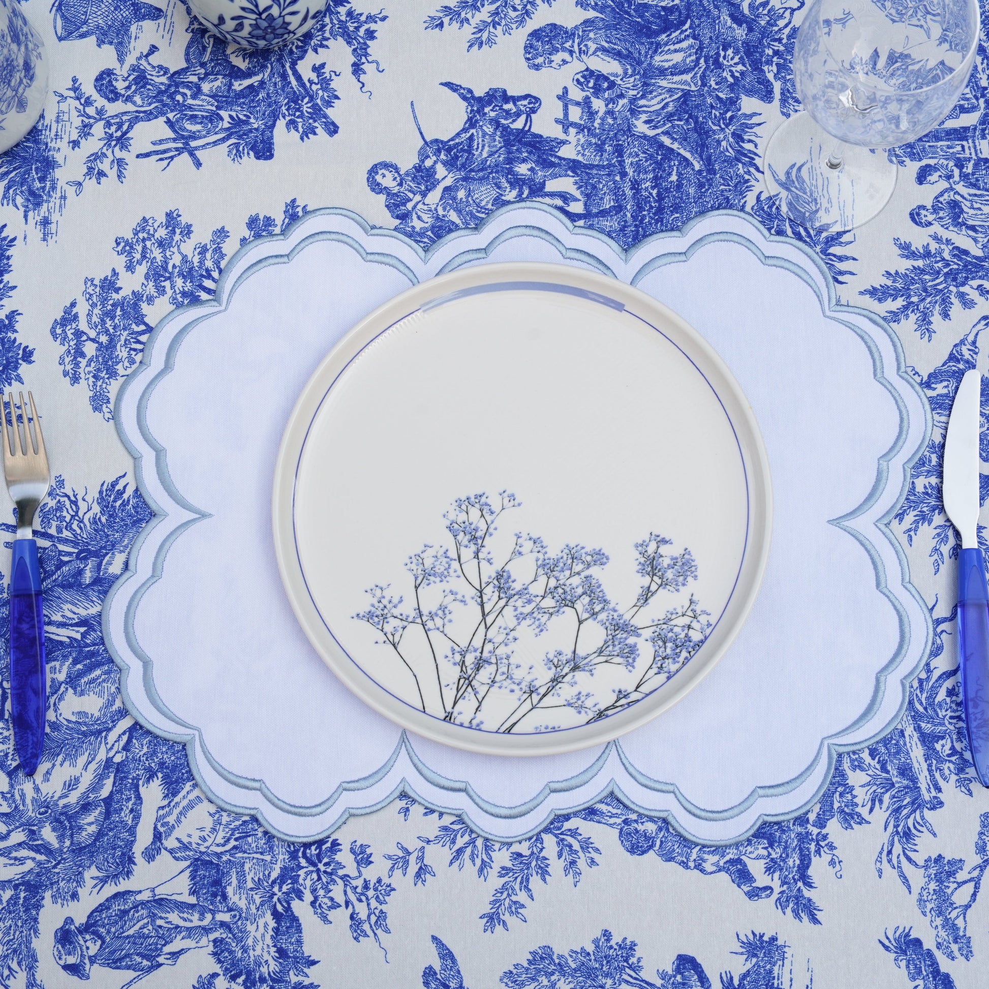 A blue and white floral patterned placemat with a round white plate and blue cutlery on top of it, displayed against a white background.
