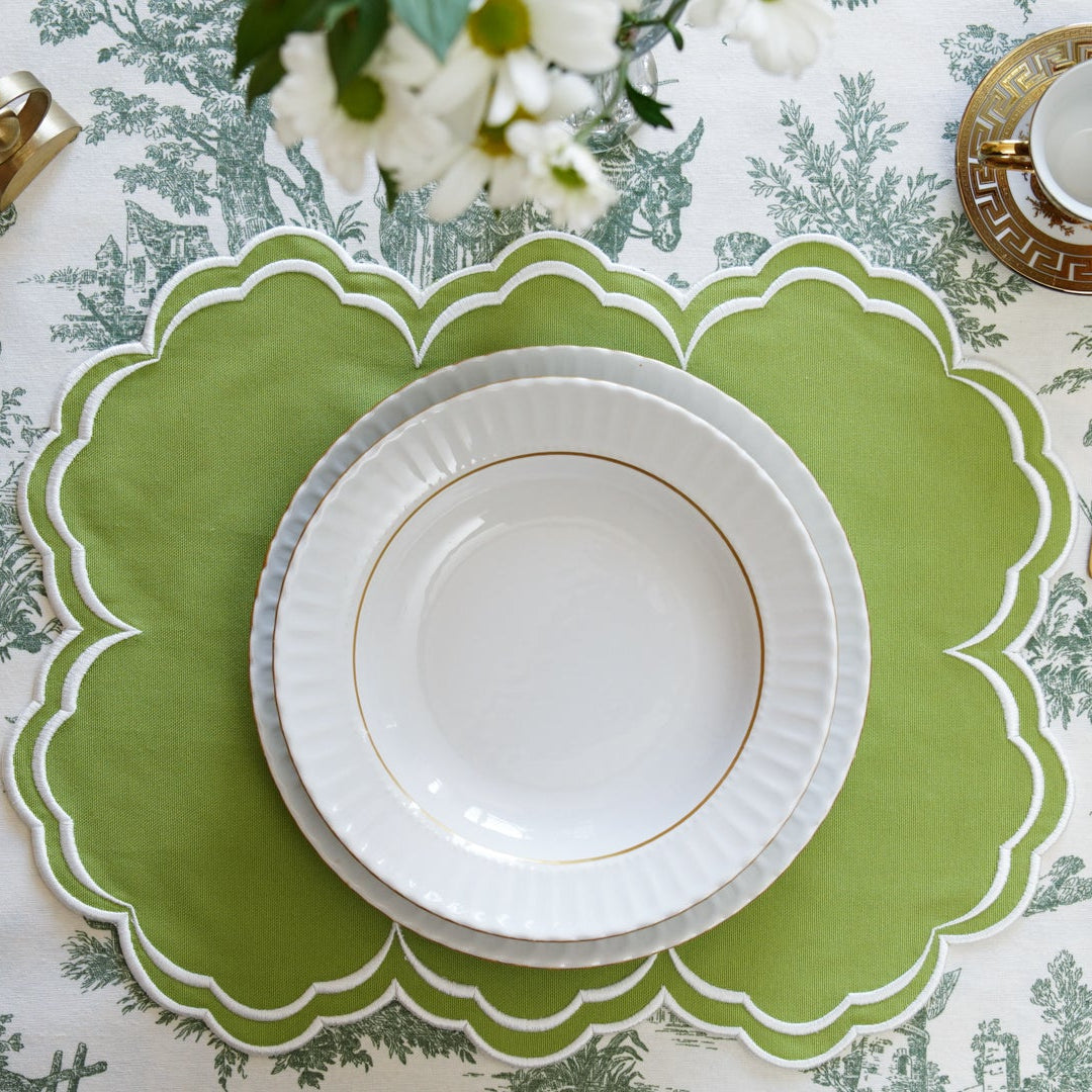 Table setting with a green placemat, white plate, gold cutlery, and floral decorations on a patterned tablecloth.
