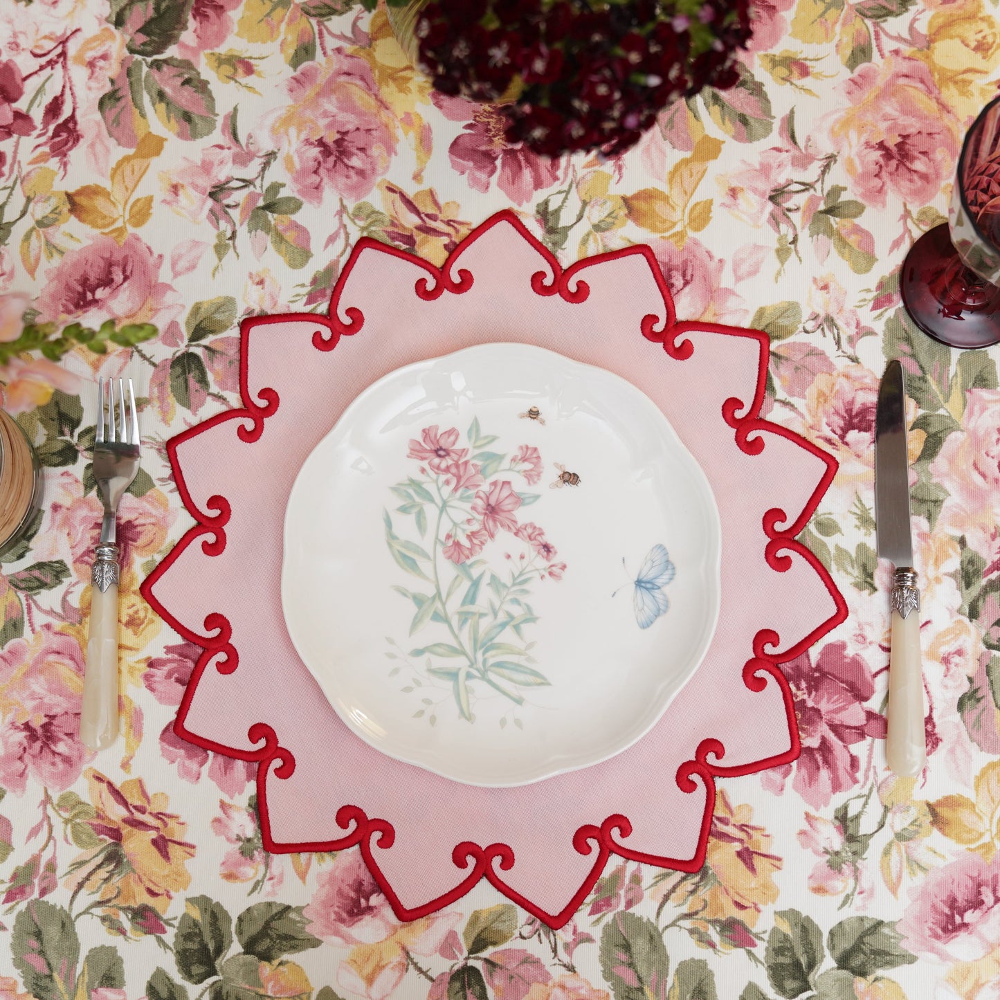 A round placemat with a pink border and red trim, featuring a floral pattern, placed on a table with a floral tablecloth.