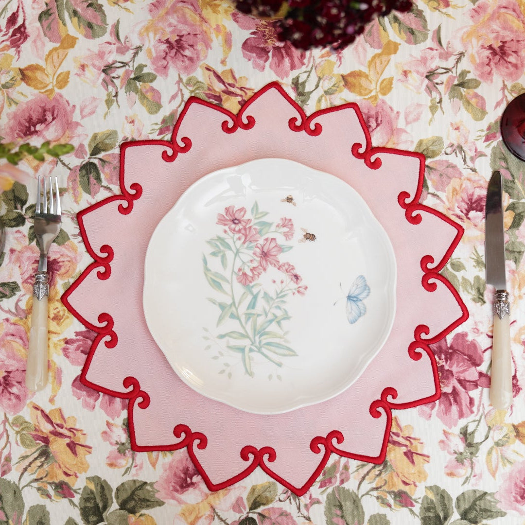 A round fabric placemat with a pink border and floral embroidery in the center, displayed on a table with a floral tablecloth.