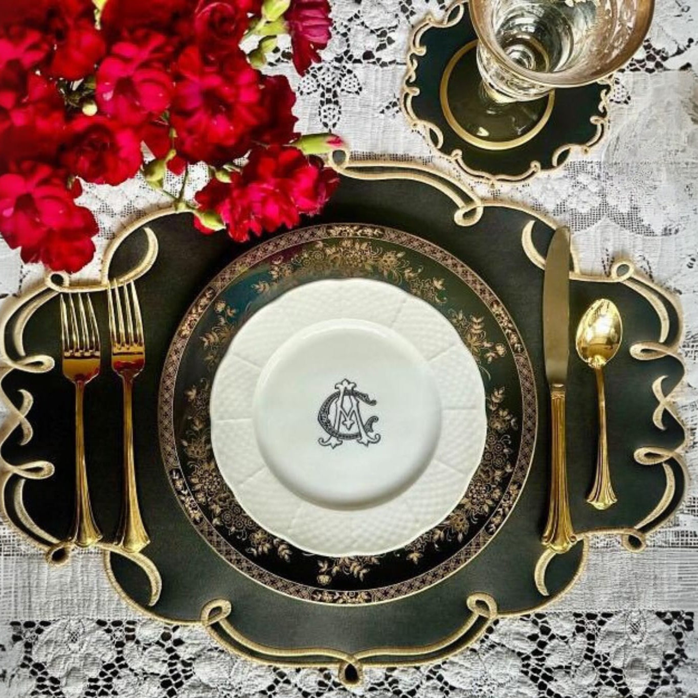 A black faux leather placemat and coaster set with gold embroidery and detailing, presented on a table with a plate, cutlery, and flowers.
