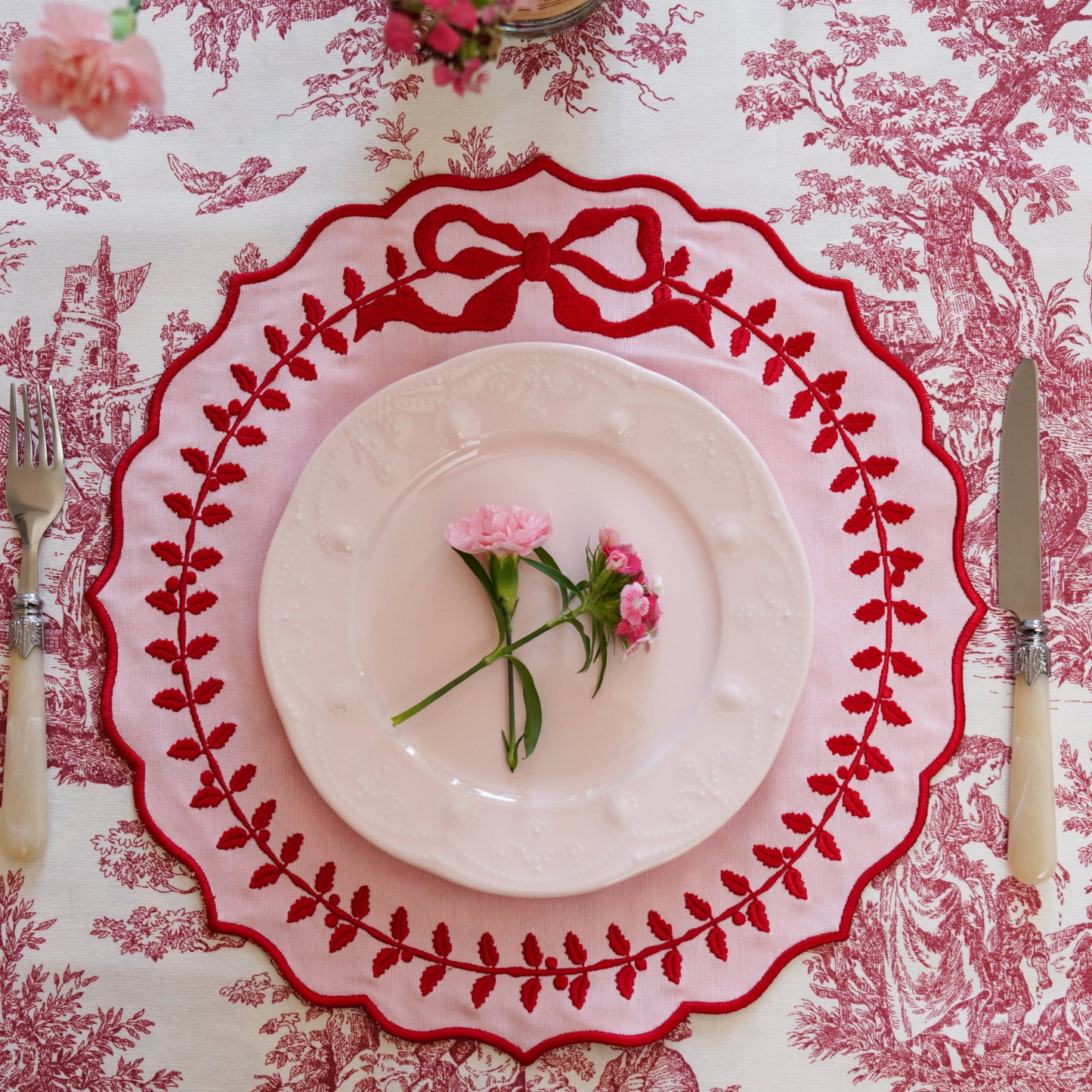 A champagne beige fabric placemat with red trim, displayed on a table with a patterned tablecloth, featuring a small pink flower in the center.
