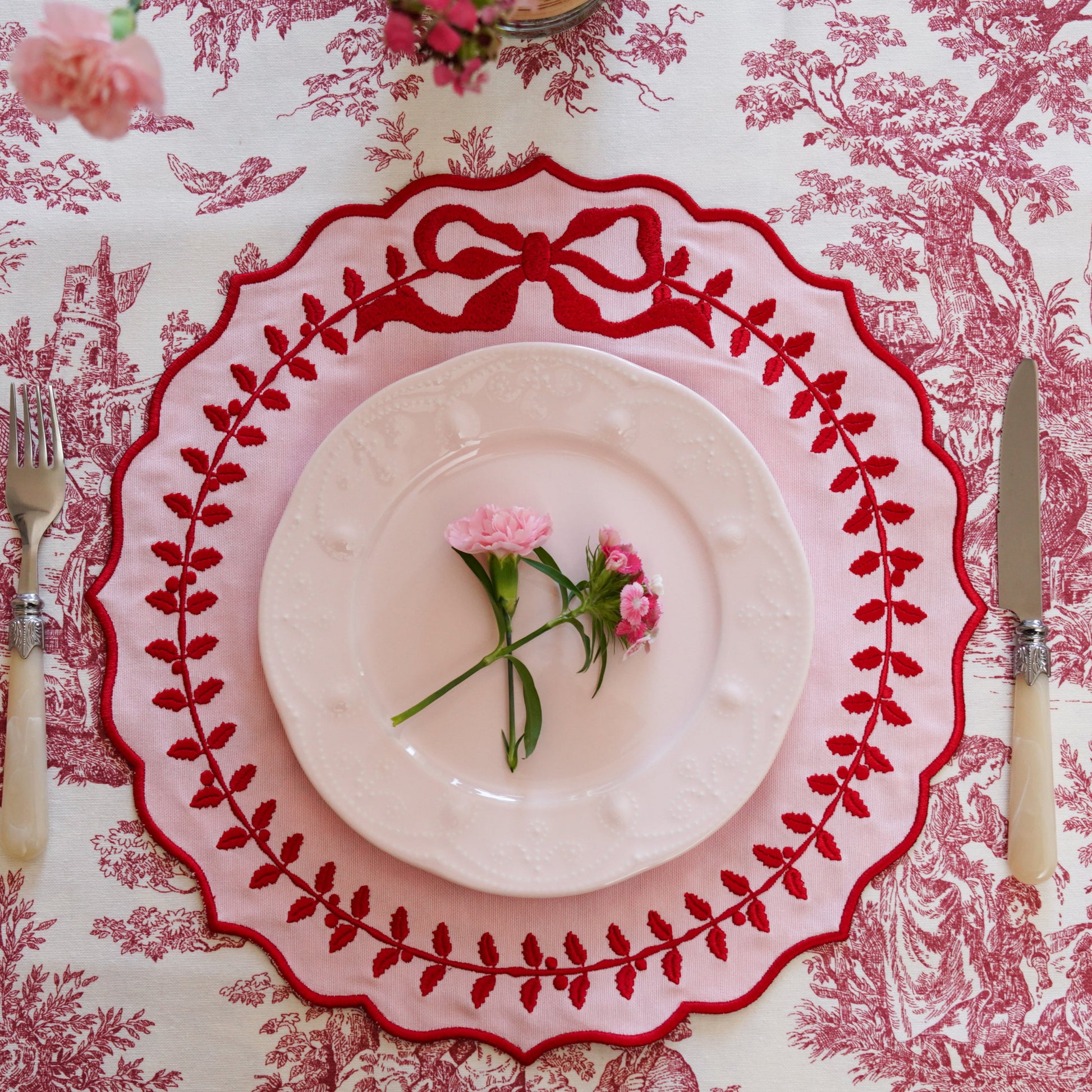A champagne beige fabric placemat with red trim, displayed on a table with a patterned tablecloth, featuring a small pink flower in the center.