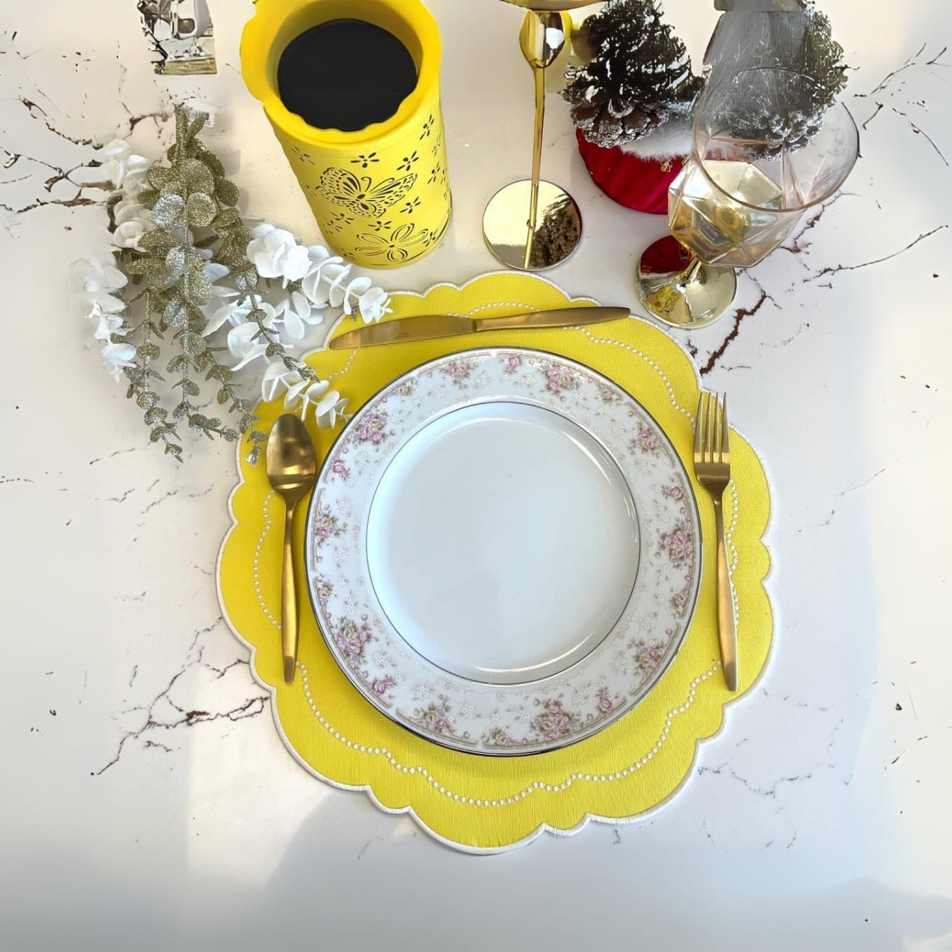 A yellow faux leather placemat is laid on a table with a white plate, silver cutlery, and a glass of red wine. The placemat has a scalloped edge design and there are decorative elements in the background suggesting a dining setting.