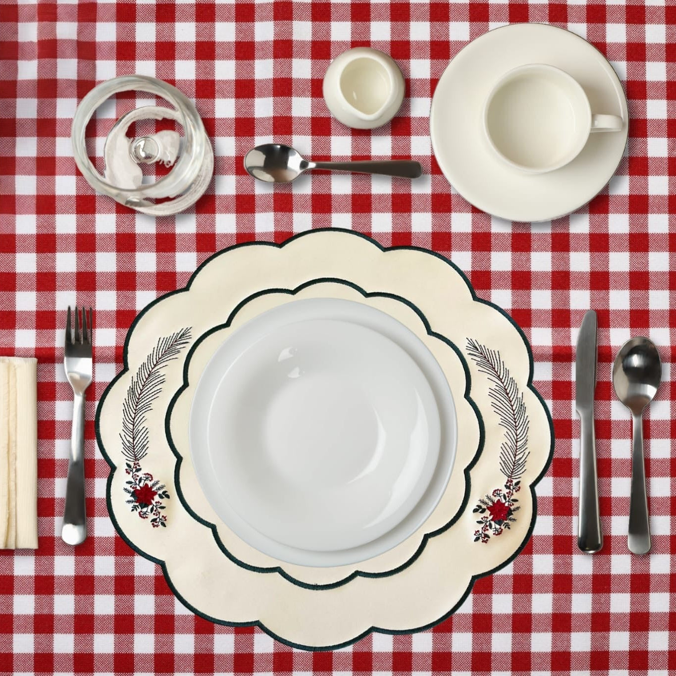 A set table with white dishes and a red and white checkered tablecloth, featuring faux leather placemats with embroidered poinsettia designs.