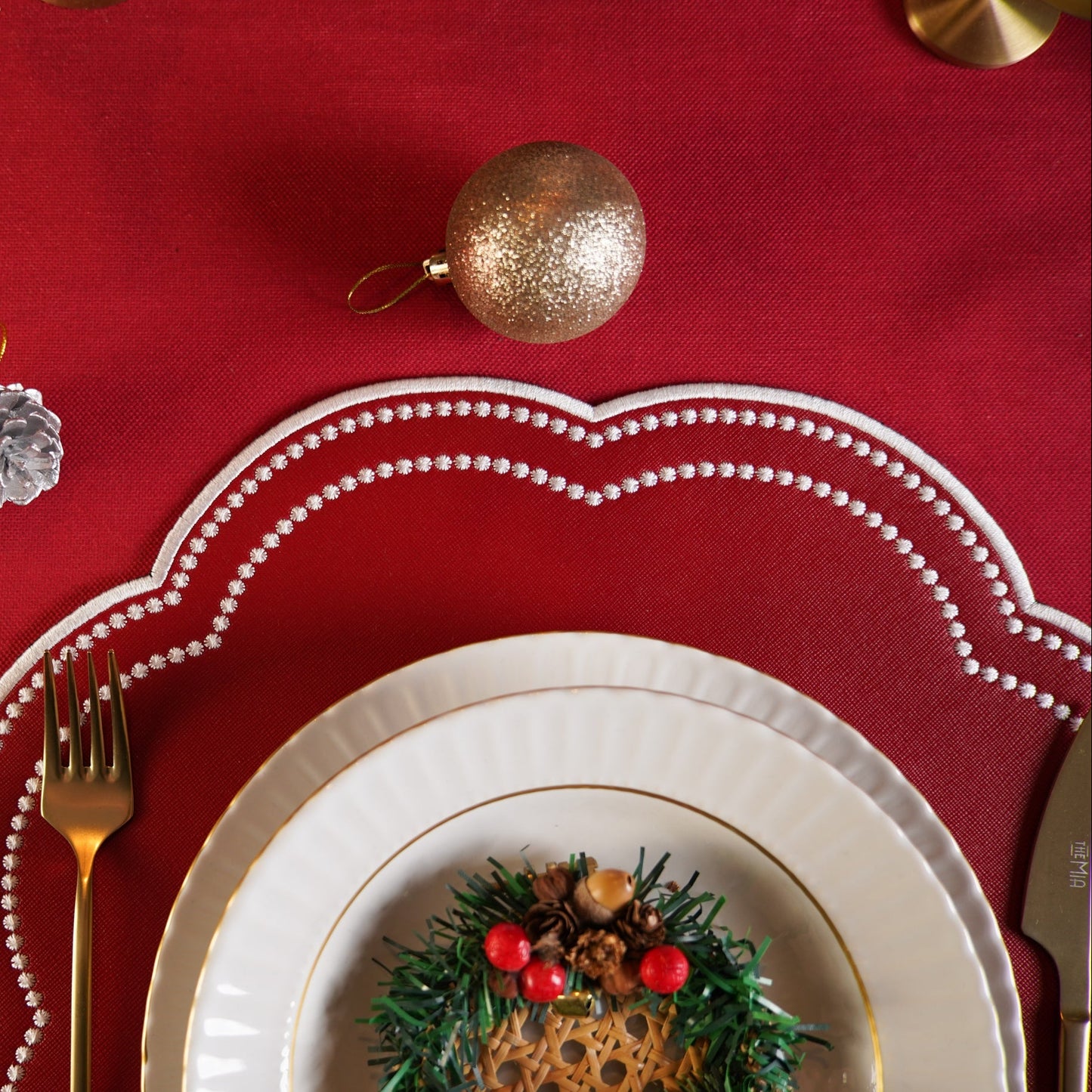 Christmas-themed table setting with red tablecloth, white plates, gold cutlery, and decorative elements.