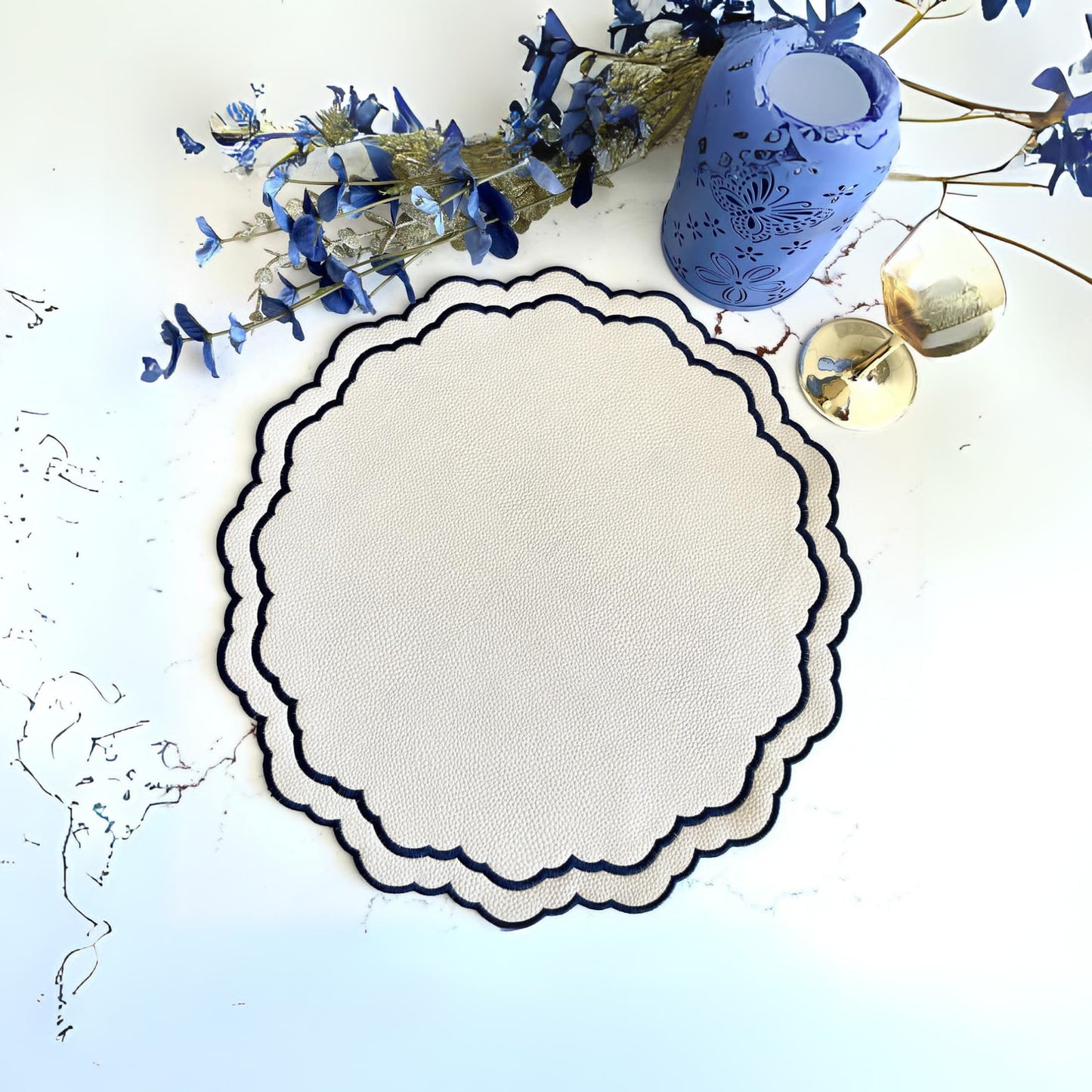 A round faux leather placemat with a decorative border in navy blue, displayed on a white surface, accompanied by a blue floral patterned napkin and some dried blue flowers.