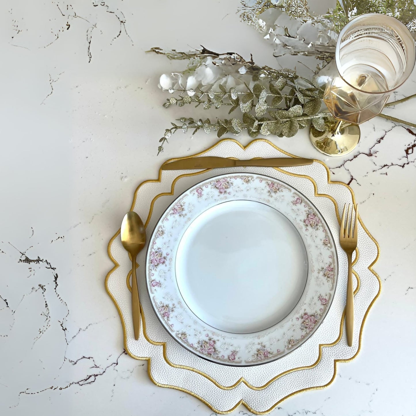 A set table featuring a white plate with gold trim, a glass of water, and a gold fork and knife. The table setting is complemented by an ivory faux leather placemat with embroidery details.