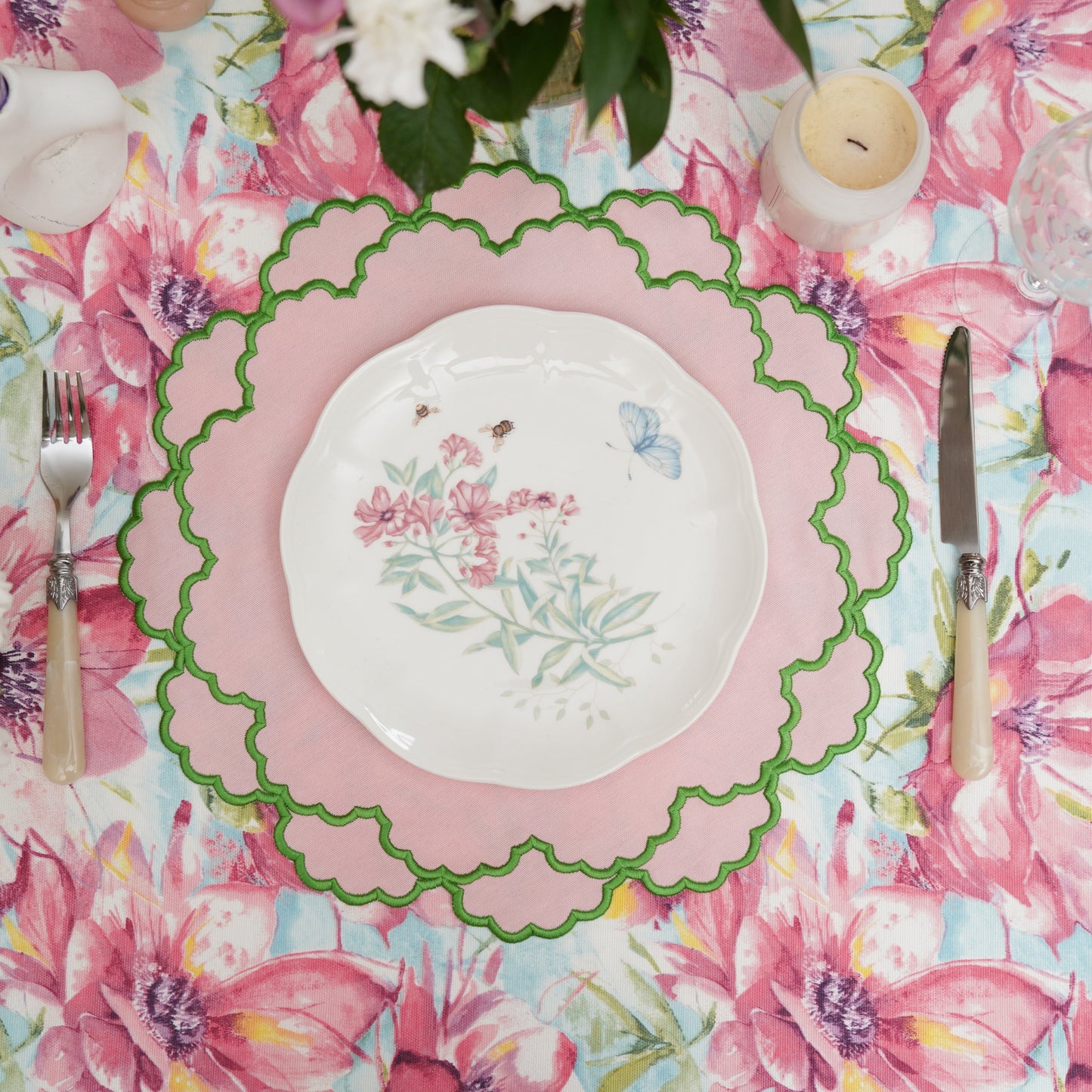 A light pink fabric placemat with green trim, displayed on a table with a floral tablecloth, a plate, and cutlery.