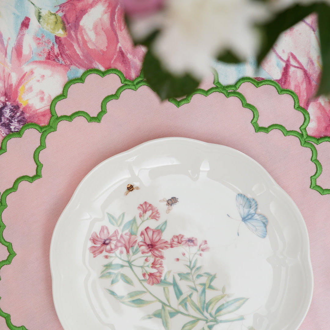 A close-up of a light pink fabric placemat with a green trim, featuring a floral pattern and an embroidered butterfly design in the center, with a fork and a plate on top of it.