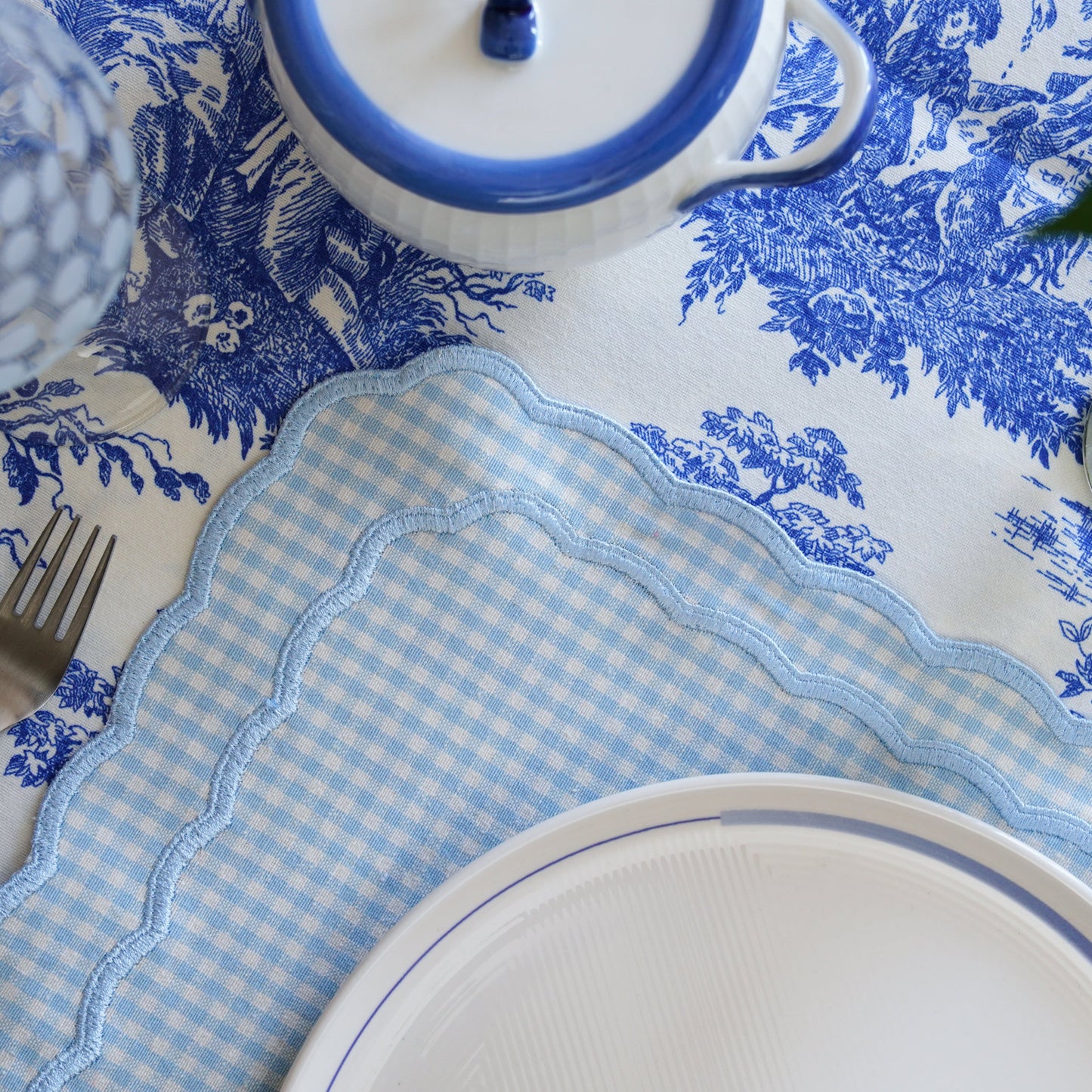 Table setting with blue and white patterned tablecloth, gingham placemat, and white plates.