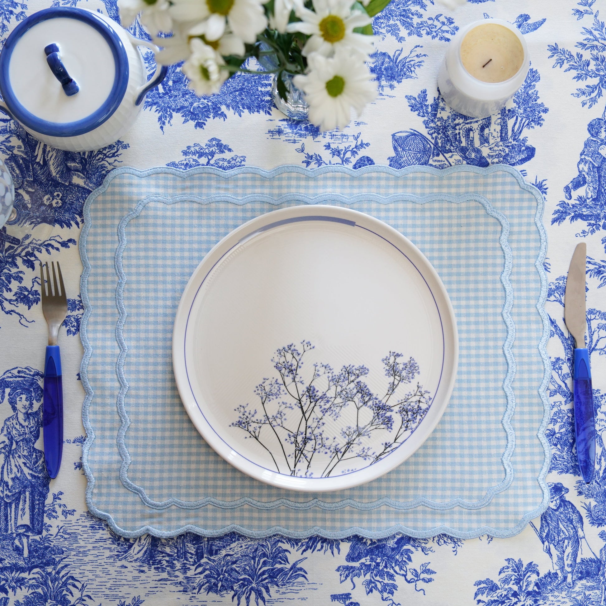 A blue and white gingham fabric placemat with floral embroidery, displayed on a dining table setting with crockery and cutlery.