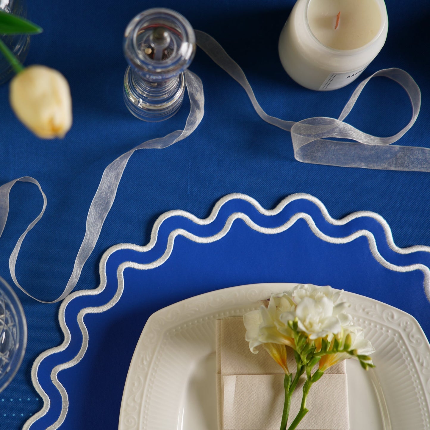 Decorative table setting with a white plate, flowers, and a candle on a blue surface.