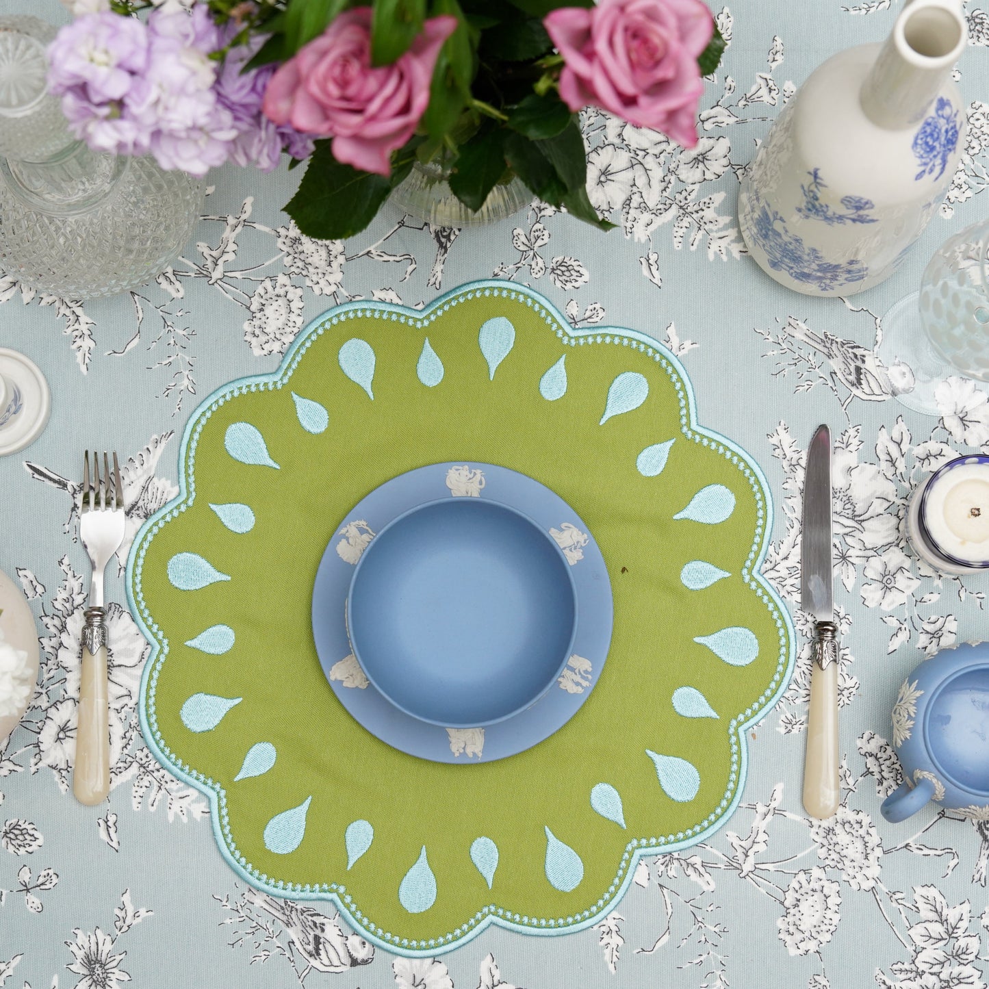 A green fabric placemat with blue trim, displayed on a table setting among other dining accessories.