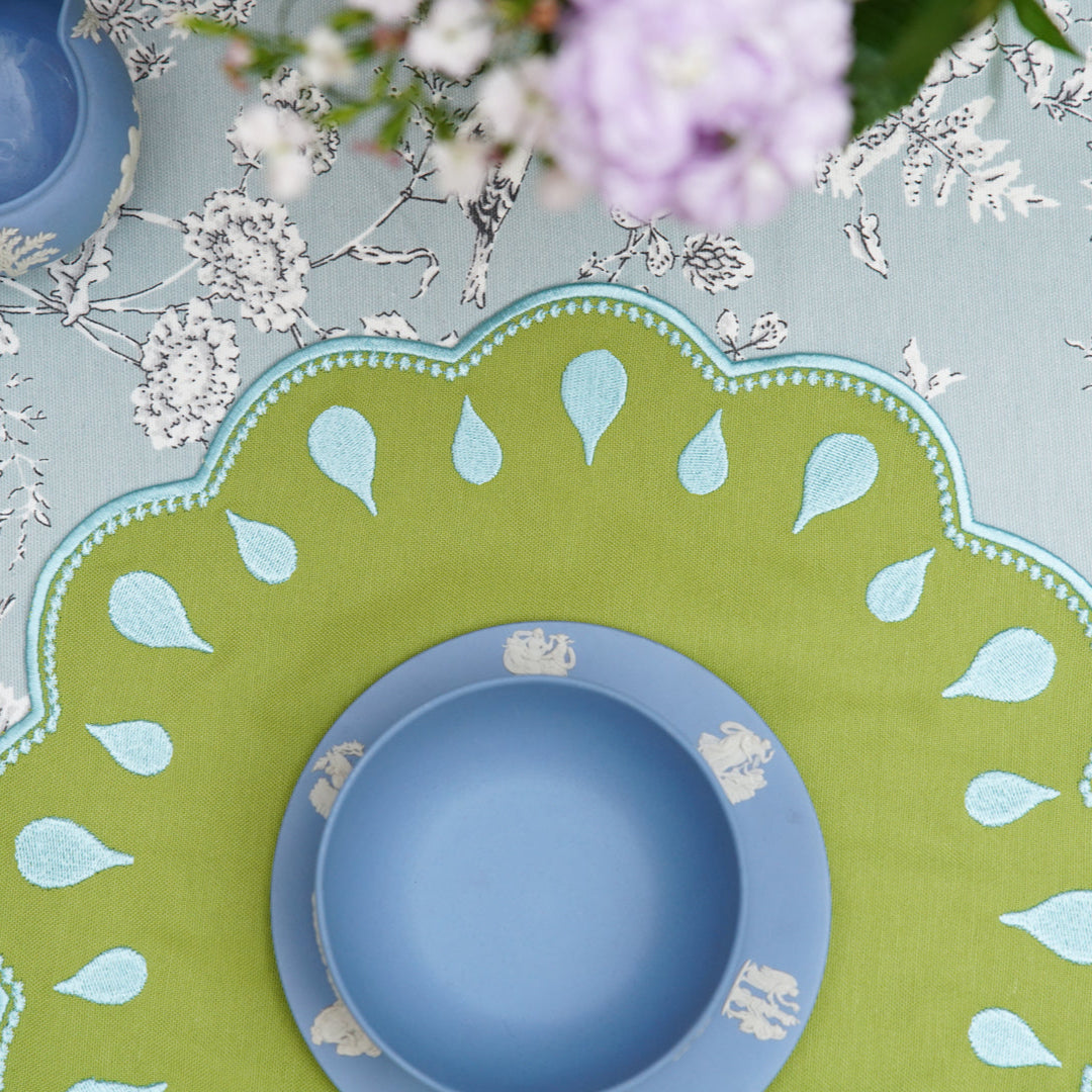A green fabric placemat with blue droplet embroidery around the edge, displayed on a table with a blue plate on top and cutlery to the side.