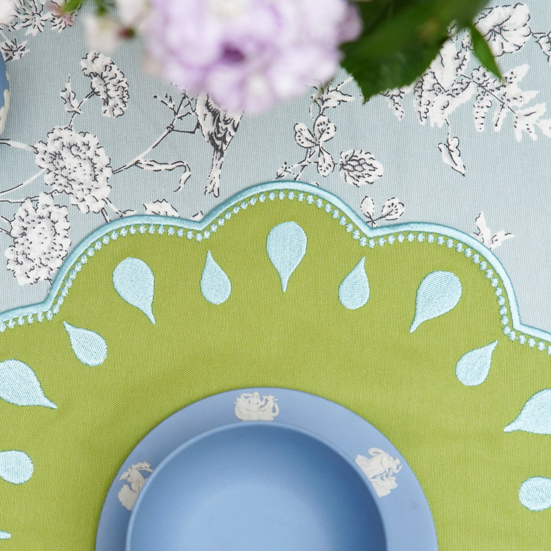 Table setting with a green and white decorative plate, blue bowl, and purple glass on a light blue floral-patterned tablecloth.