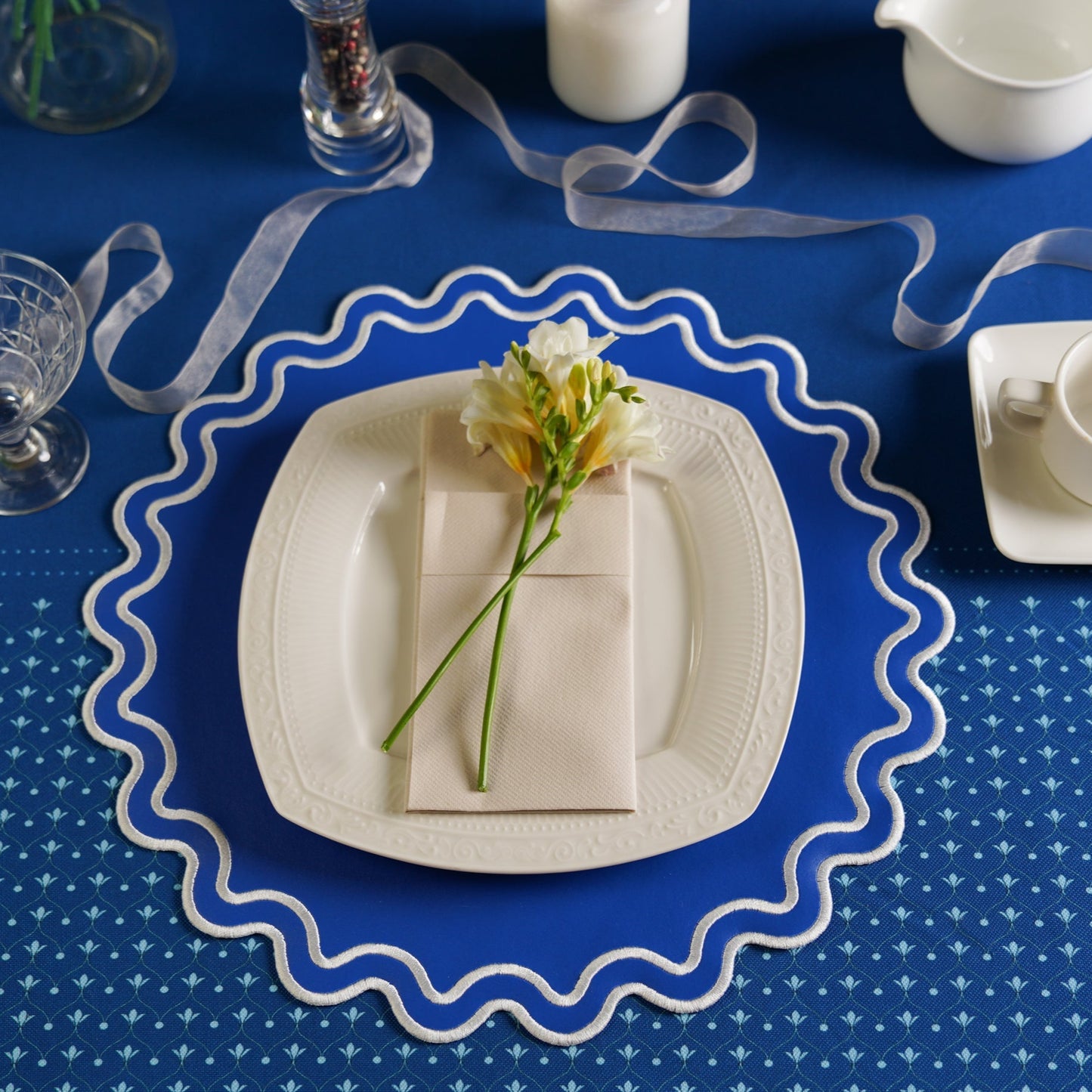 Table setting with a white plate, napkin, and flower on a blue tablecloth with decorative elements.