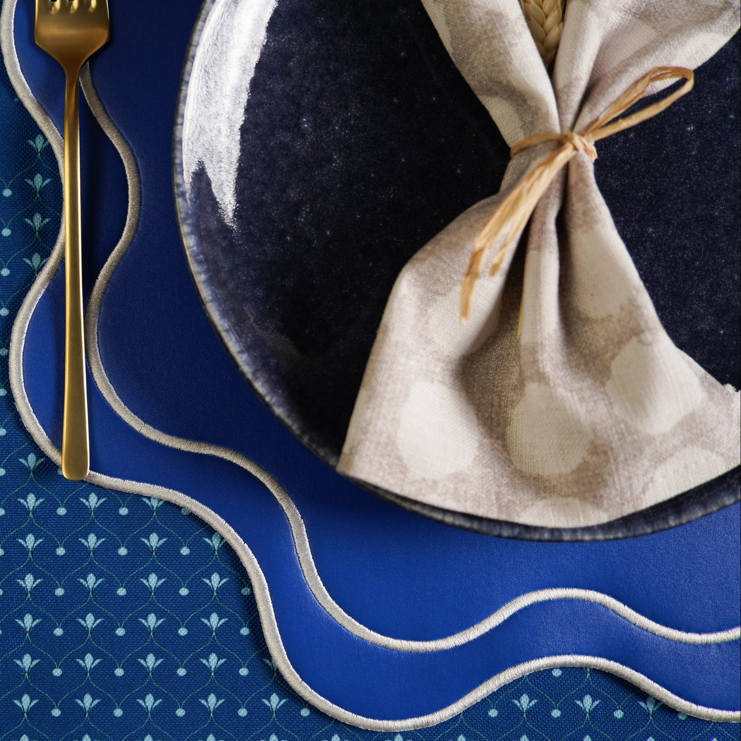 Napkin tied on a blue plate with a gold fork on a matching blue placemat.
