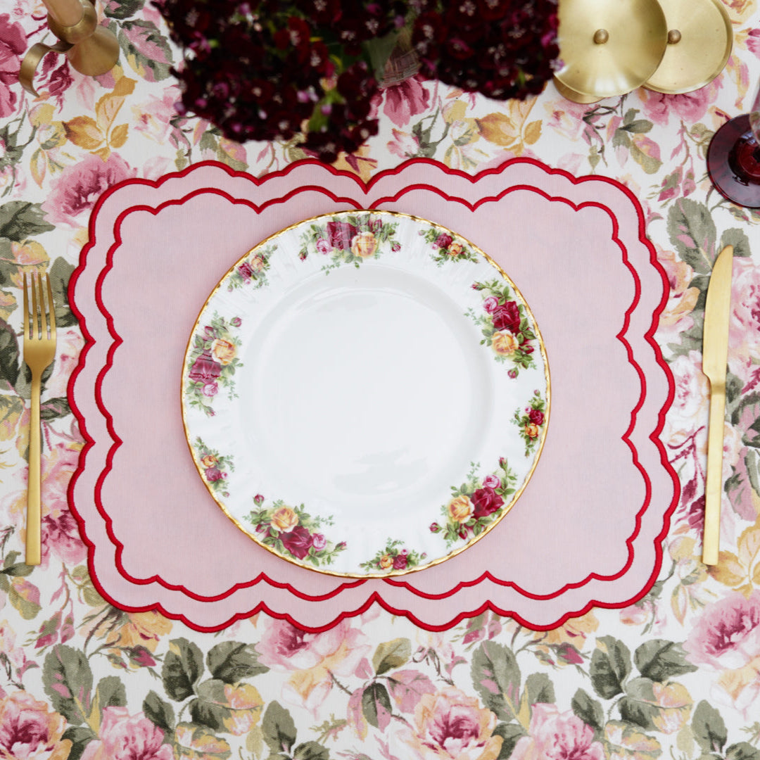 A round placemat with a pink floral pattern and red trim, displayed on a table with a dish and cutlery.
