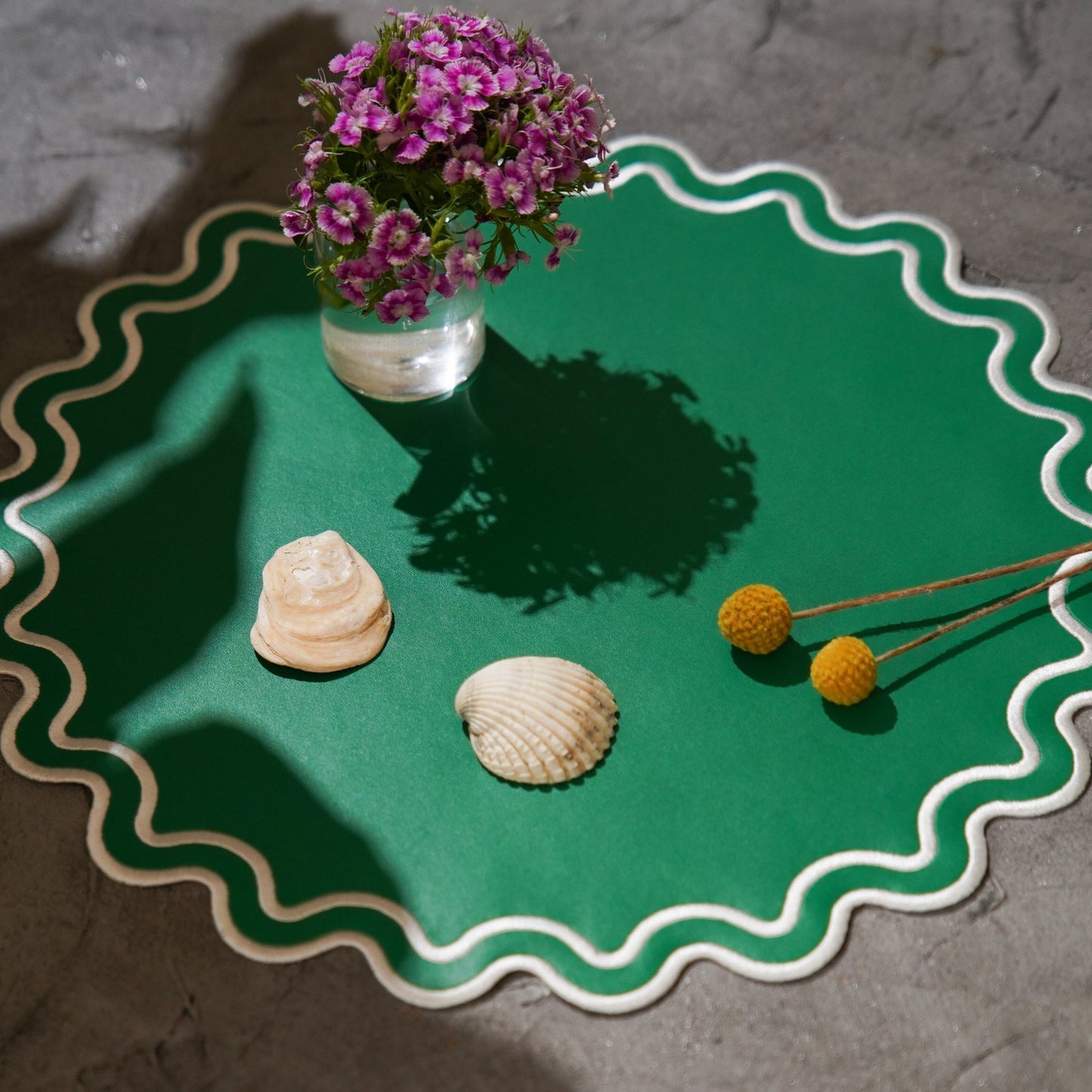 Green ceramic plate with a vase of purple flowers, two seashells, and yellow flowers on a gray surface.