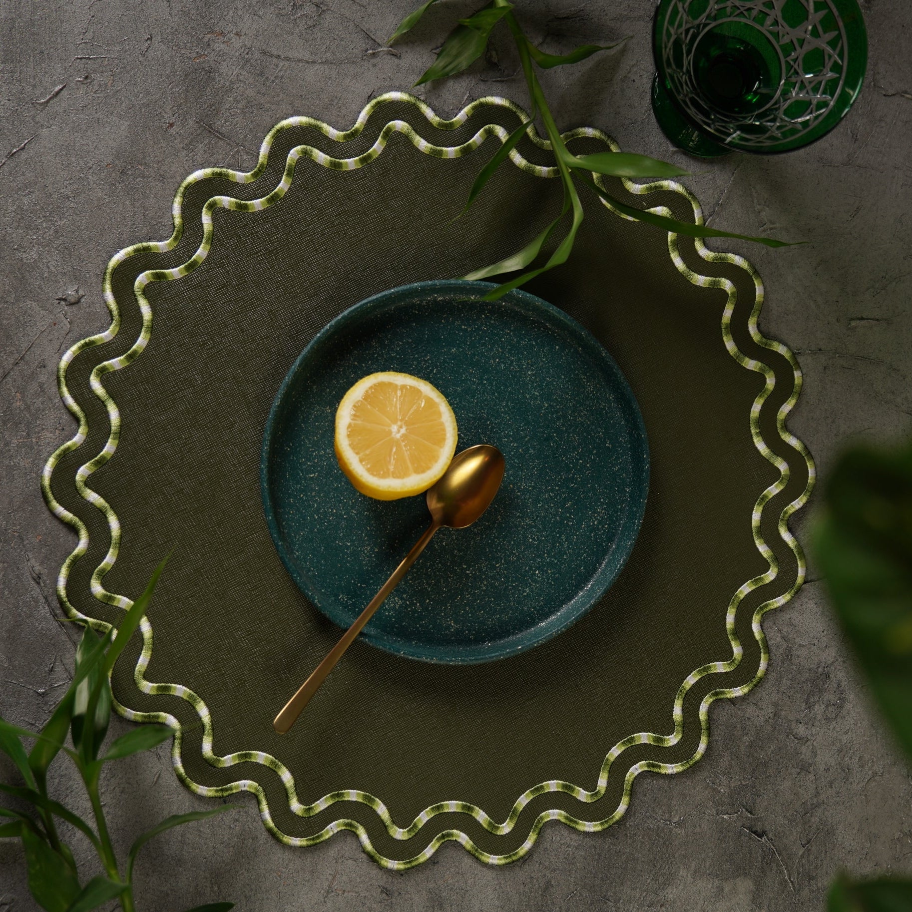 A dark green faux leather placemat with a decorative edge, displayed on a table with a plate and lemon slice on top, surrounded by green foliage.
