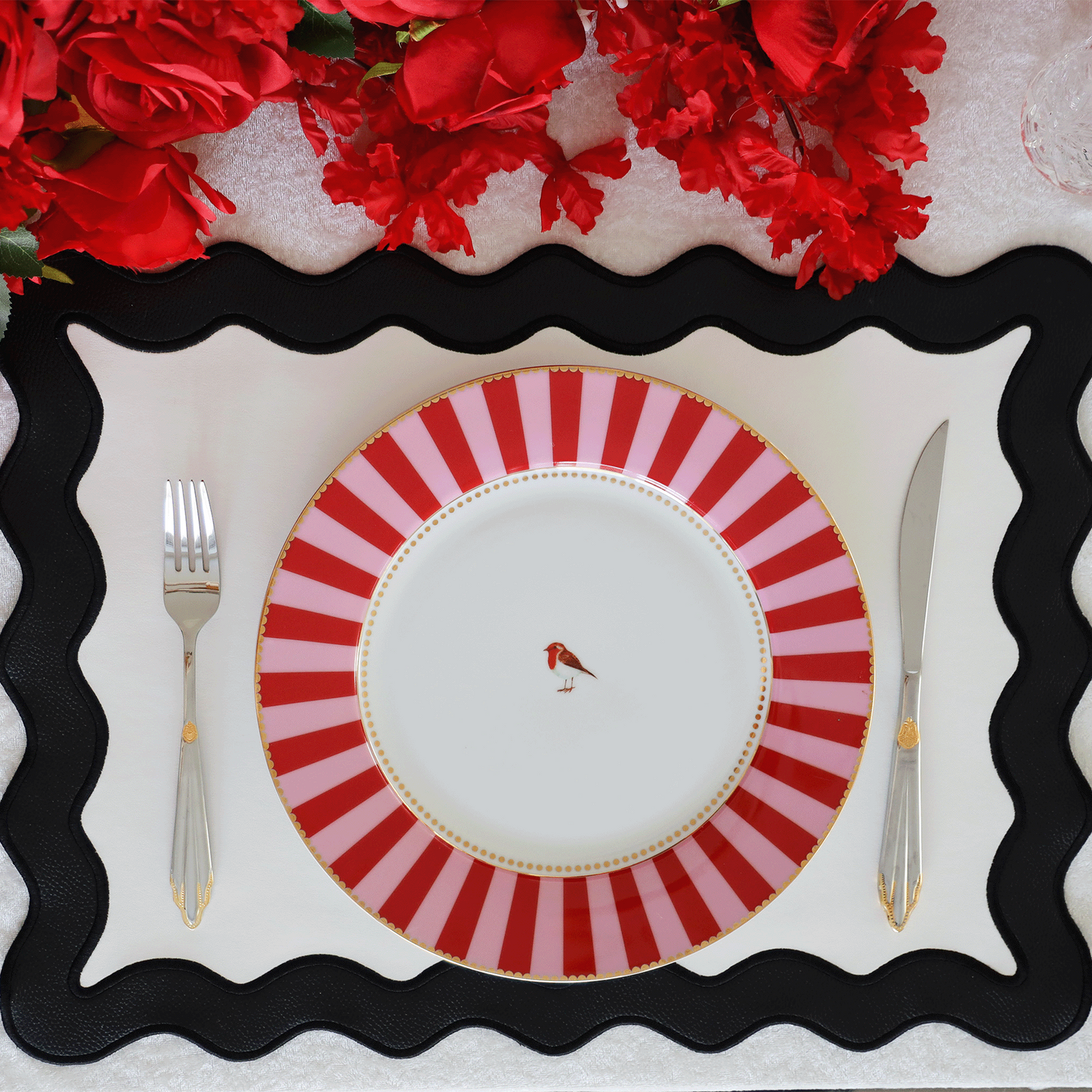 A black and white faux leather placemat with a decorative edge, presented on a table with a red and white striped plate on top and red flowers in the background.