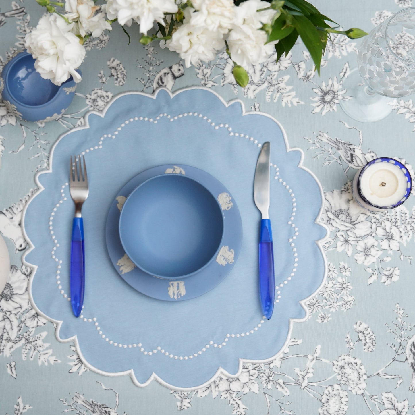 A round blue fabric placemat with white trim, displayed on a table with cutlery, flowers, and a candle.