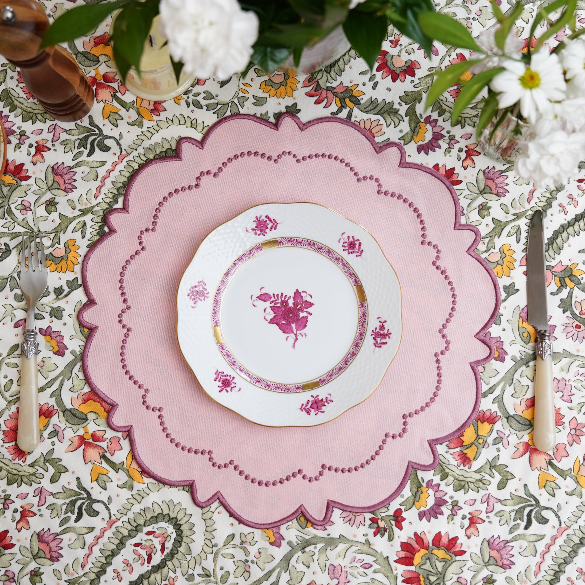 A pink fabric placemat with floral embroidery is displayed on a table with a patterned tablecloth and a white plate on top.