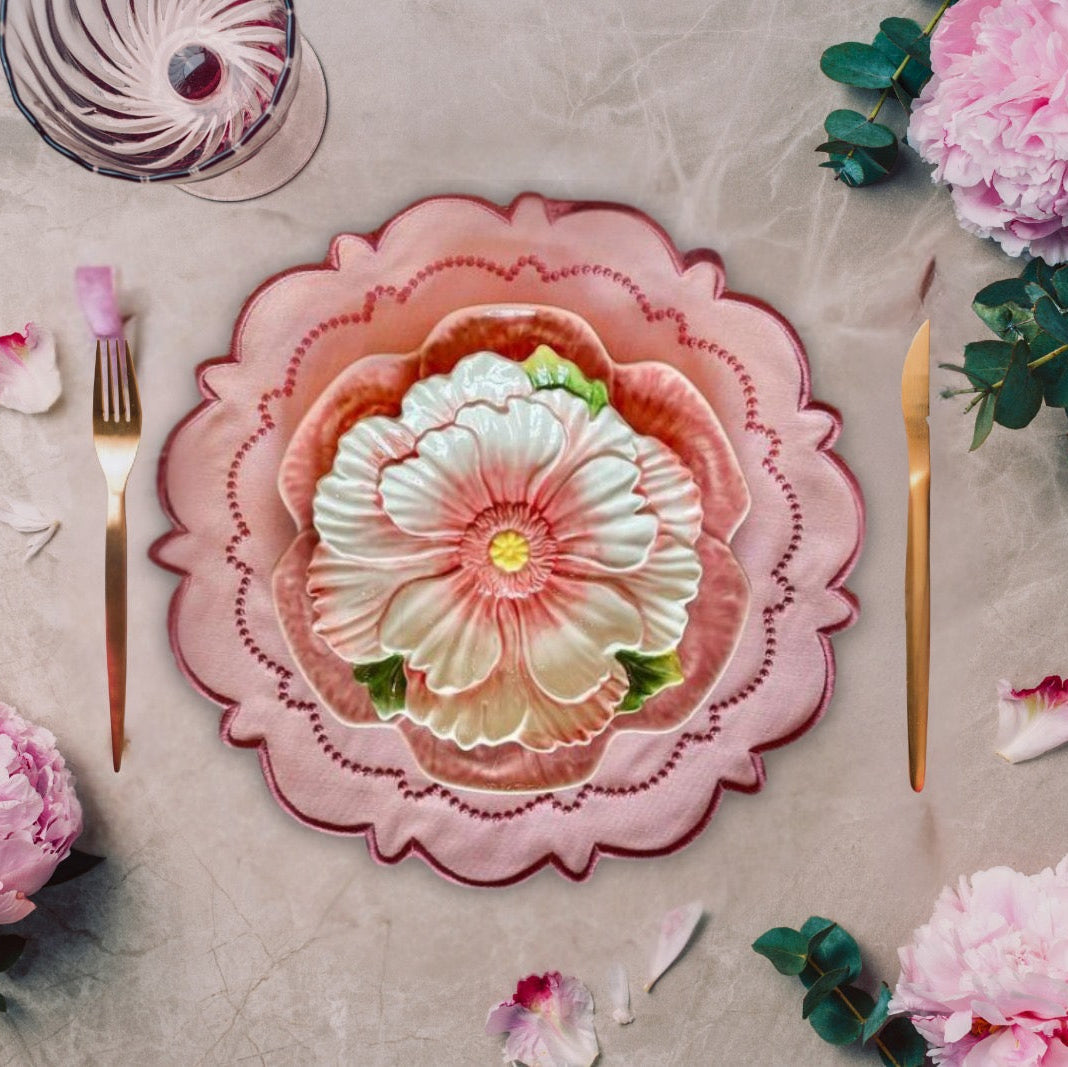 A pink fabric placemat set on a table, with a decorative floral design in the center. Surrounding the placemat are pink flowers and cutlery.
