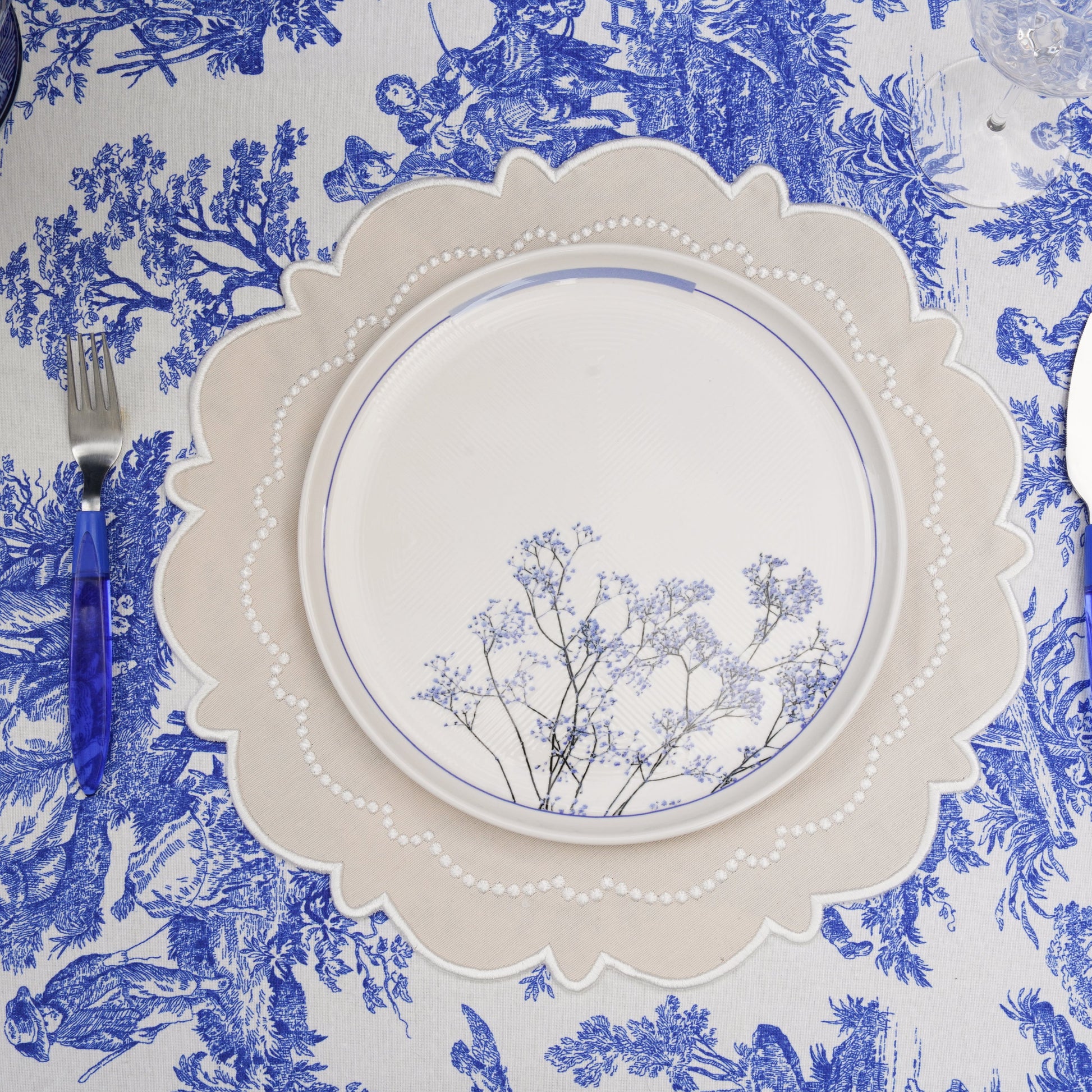 A round, cream-colored fabric placemat with intricate white embroidery, displayed on a table with a blue floral patterned tablecloth, accompanied by a white plate and blue utensils.