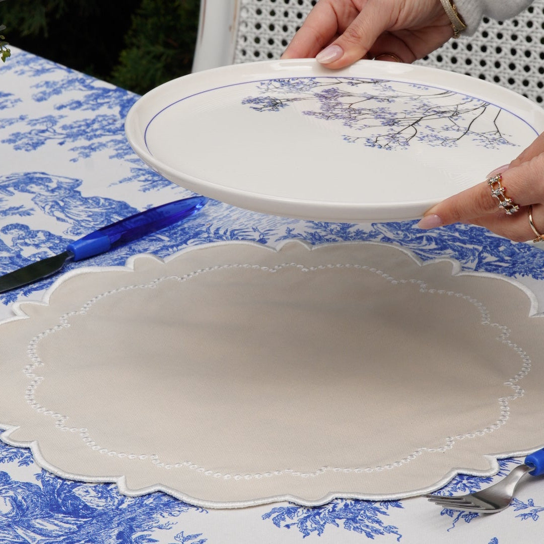 A person is setting a table with a round, cream-colored fabric placemat featuring intricate embroidery, placed on a blue patterned tablecloth.