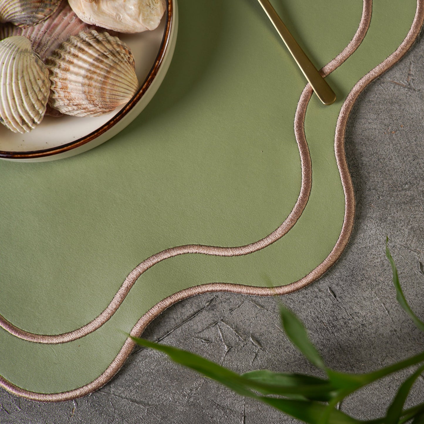 Green placemat with wavy brown border on a stone surface with shells and a spoon.