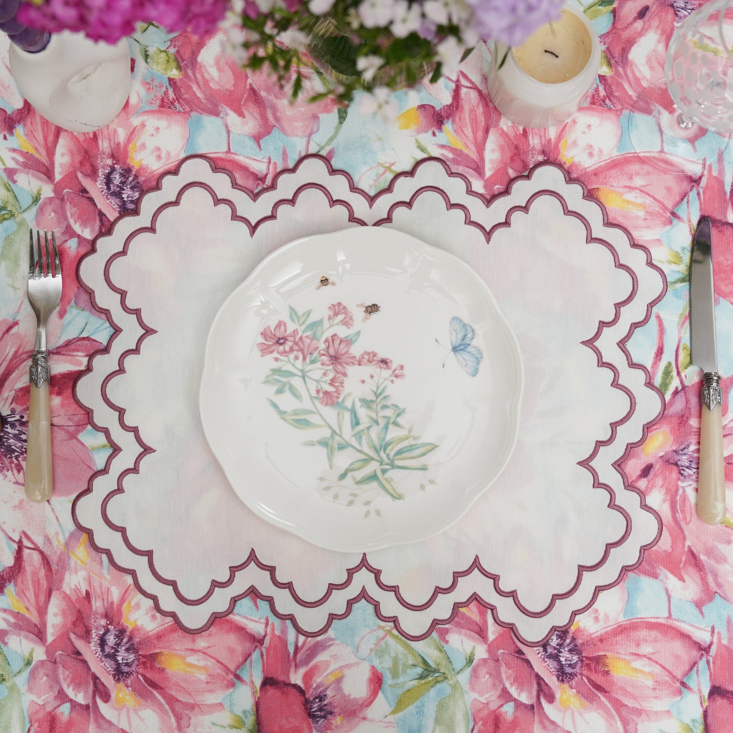 A round placemat with pink trim and floral embroidery, displayed on a table with a floral patterned tablecloth.