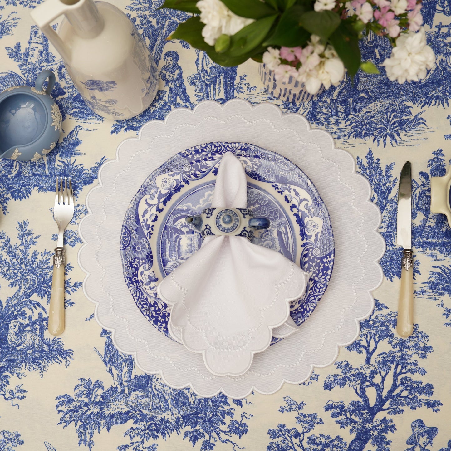 A set of white fabric placemats with embroidered detailing, displayed on a dining table setting with blue and white tablecloth.