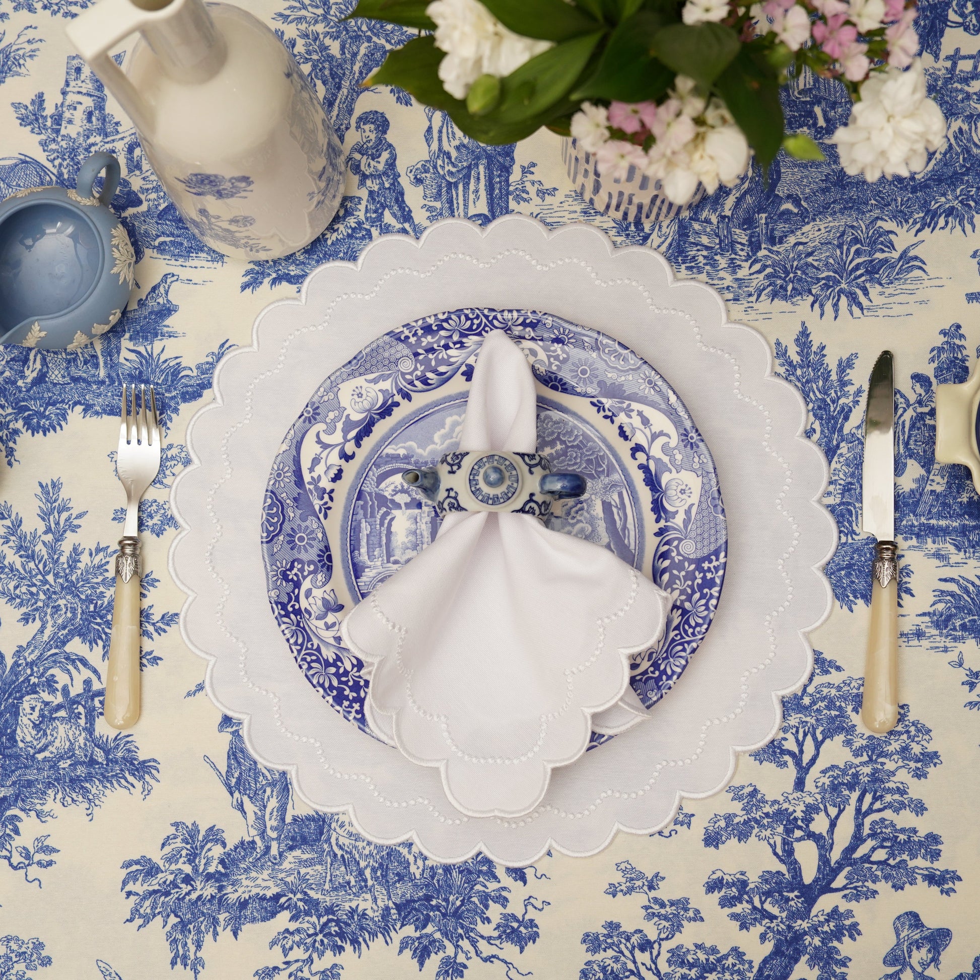 A set of white fabric placemats with embroidered detailing, displayed on a dining table setting with blue and white tablecloth.