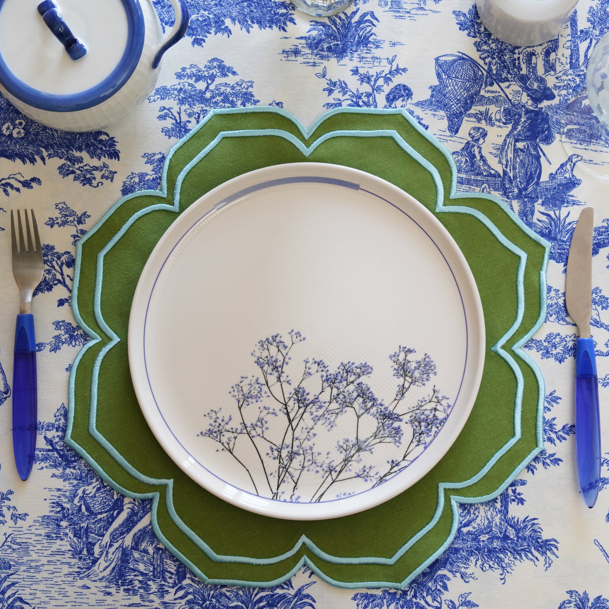 A round placemat with green and blue floral embroidery around the edge, displayed on a table with a blue and white patterned tablecloth.