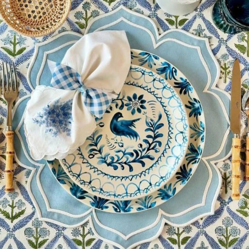 A set of light blue fabric placemats with a floral pattern, displayed on a dining table with a plate and cutlery set on top.