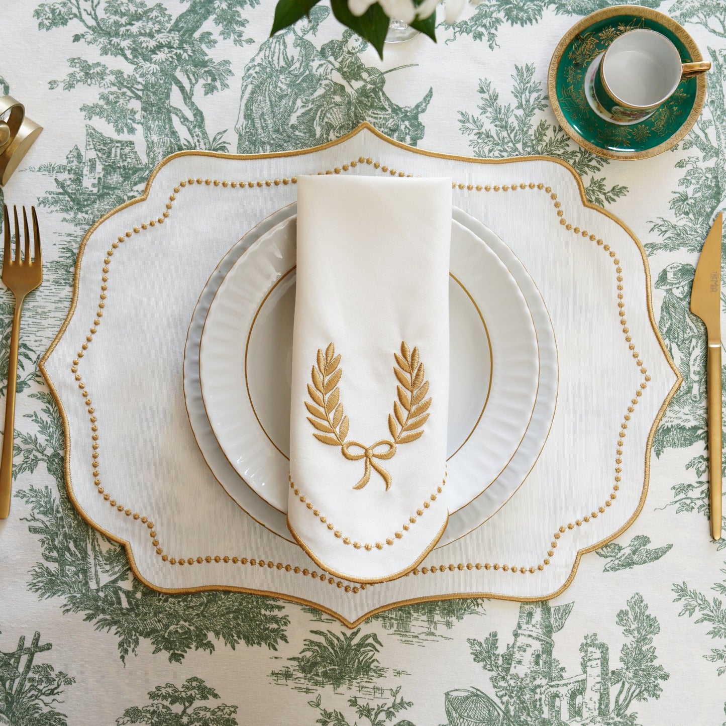 A set of embroidered fabric placemats in ivory with gold trim, displayed on a dining table setting with cutlery and a patterned tablecloth.