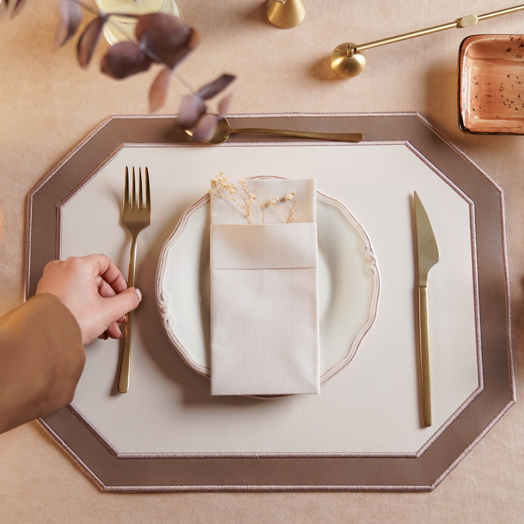 Dining table setting with a hand holding a fork, plate, and folded napkin on a decorative placemat.