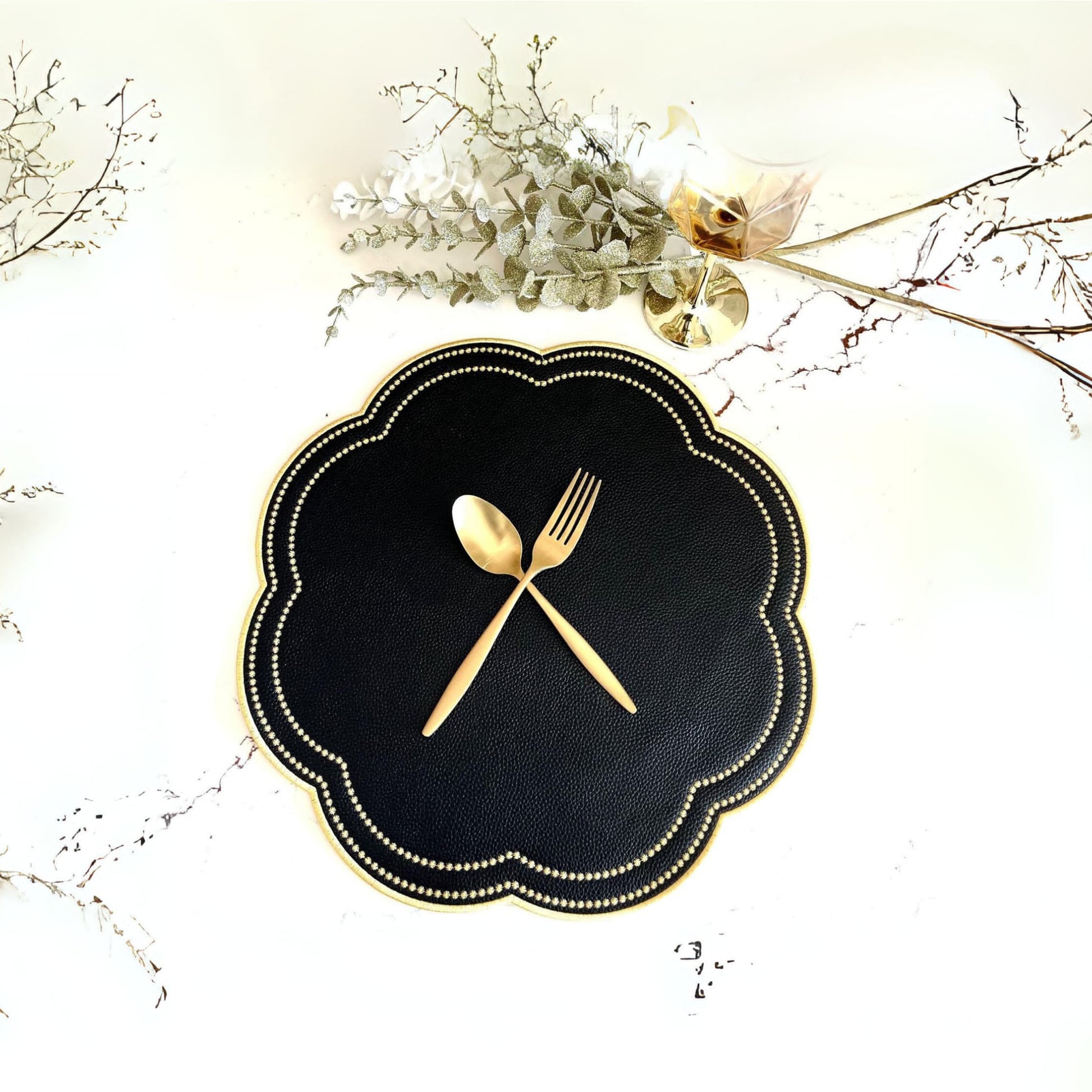 A round black faux leather placemat with gold accents and embroidery, displayed on a table with decorative foliage around it.