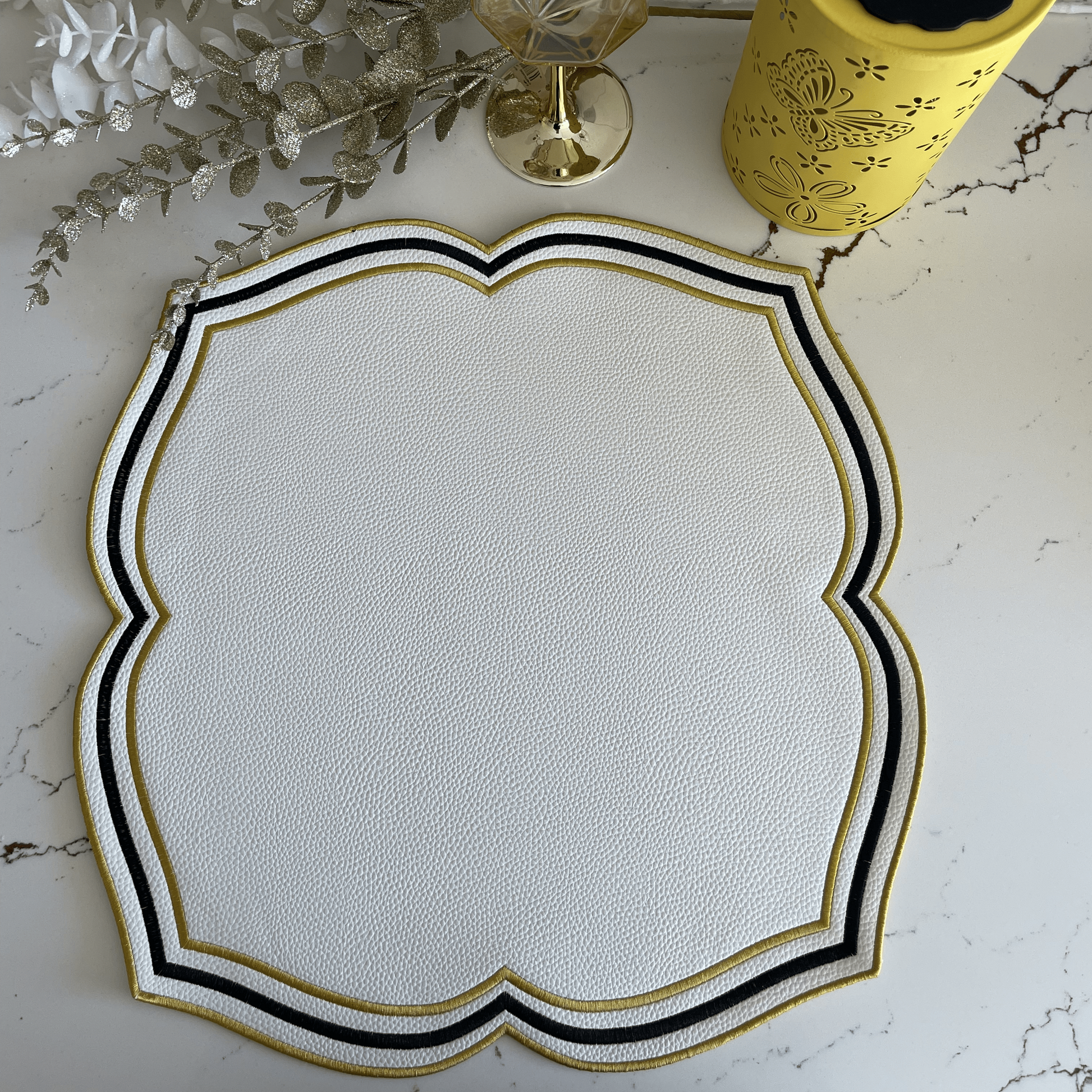 White placemat with black and gold border on a marble surface with a glass of water and decorative elements.
