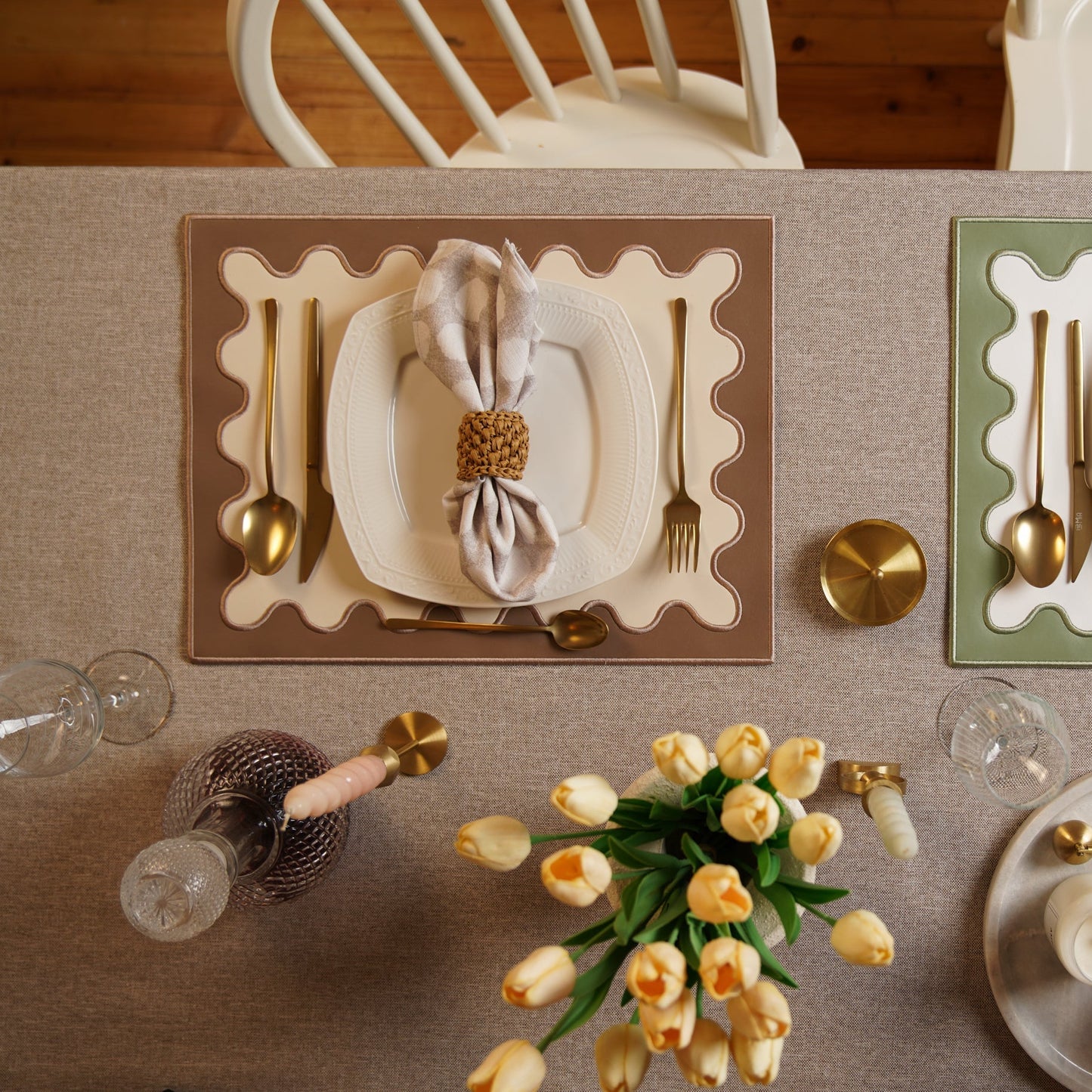 Elegant table setting with gold cutlery, white napkin, and yellow tulips on a beige tablecloth.