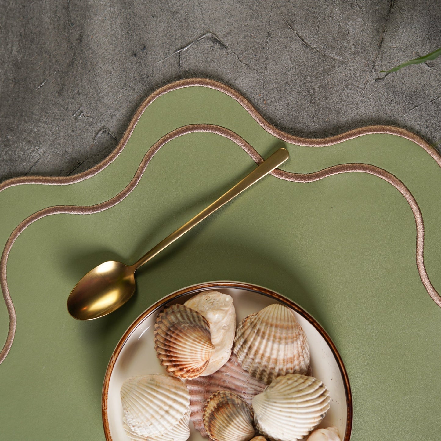 Gold spoon on a green plate with seashells against a stone background