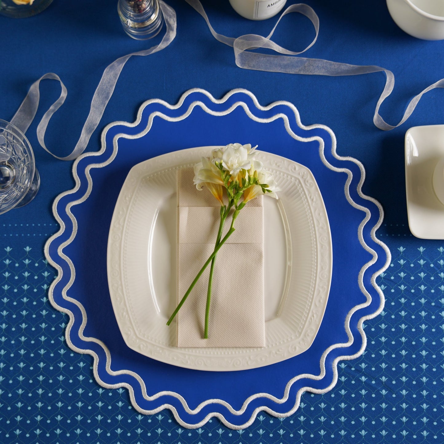 White plate with a folded napkin and flowers on a blue tablecloth with decorative ribbons.