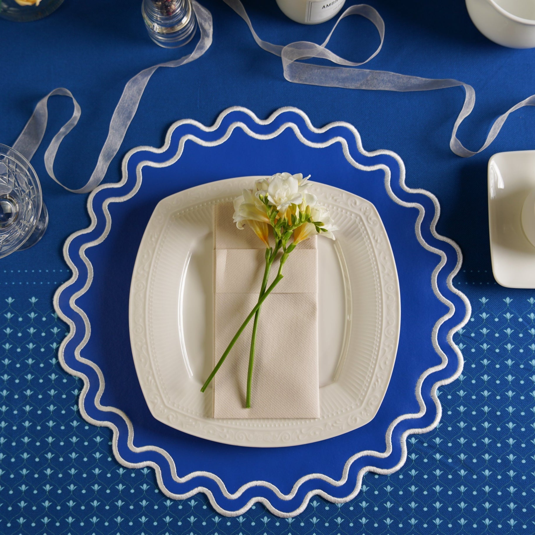 White plate with a folded napkin and flowers on a blue tablecloth with decorative ribbons.