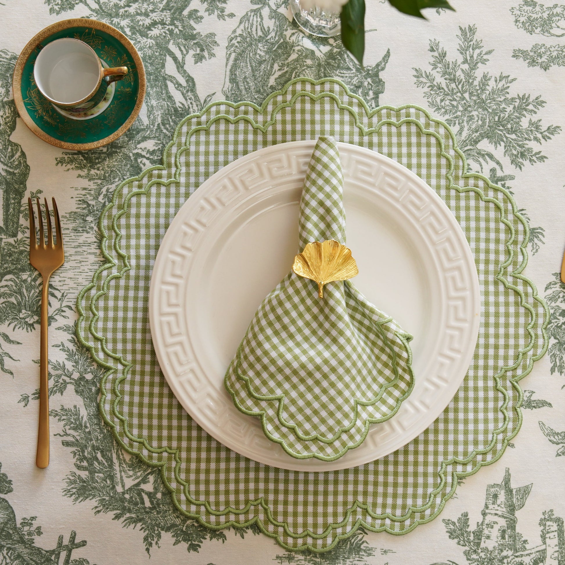 A set of green and white gingham pattern placemats with a napkin, plate, and cutlery on top, displayed on a table with a floral tablecloth.