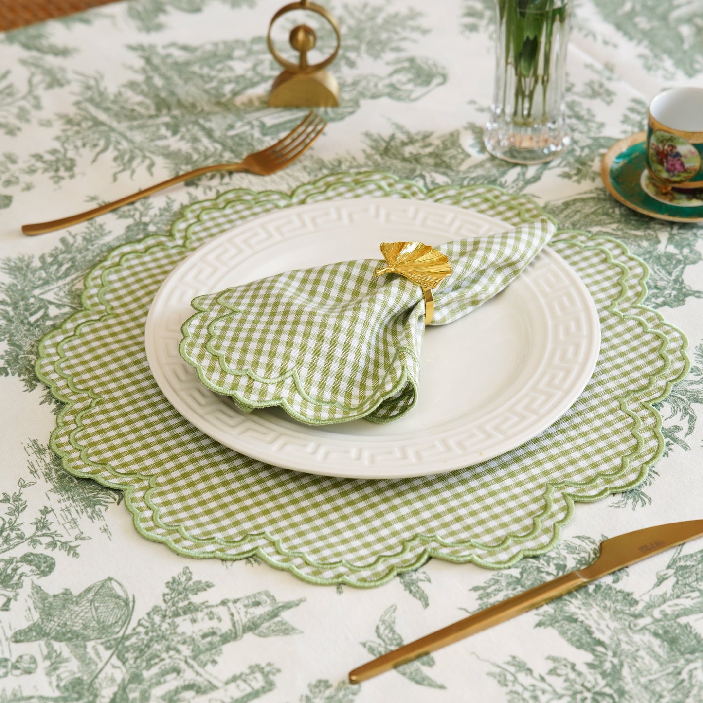 Table setting with green and white checkered napkin, white plate, and gold cutlery on a patterned tablecloth.