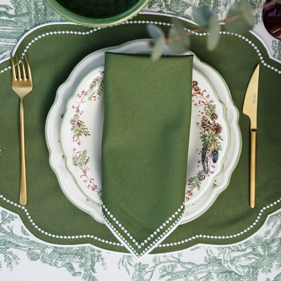 A set of green fabric placemats with white trim, displayed on a dining table, with a plate, cutlery, and a floral pattern visible.